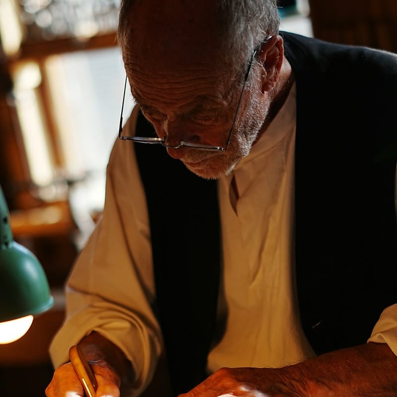 An older man in glasses and a dark waistcoat writes with a pen at his workbench, lit by a green desk lamp.