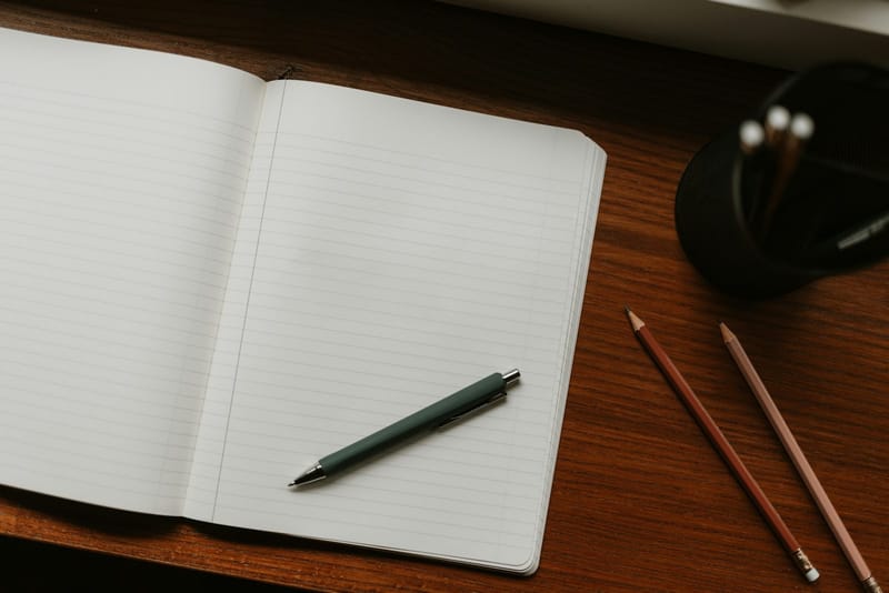 An open lined notebook with a green pen on a dark wooden desk, beside two pencils and a black cup of more pencils.