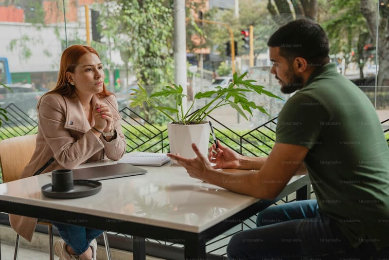 A woman listens closely as a man gestures while explaining something across a cafe table, a notebook between them.