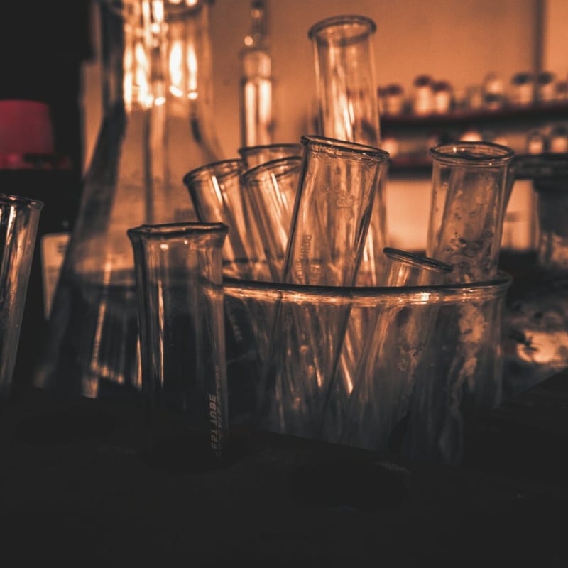 Empty laboratory flasks and test tubes standing in a beaker on a bench, backlit by warm amber light in a dim lab.
