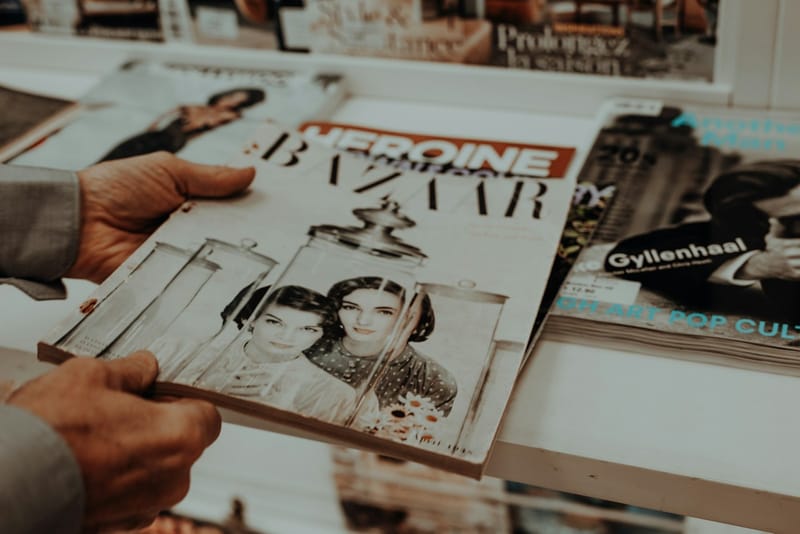 Two hands holding a printed fashion magazine above a newsstand shelf stacked with other magazines.