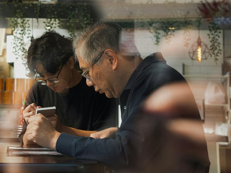 An older man and a younger one in glasses bent together over a small notebook at a café counter, seen through the window.