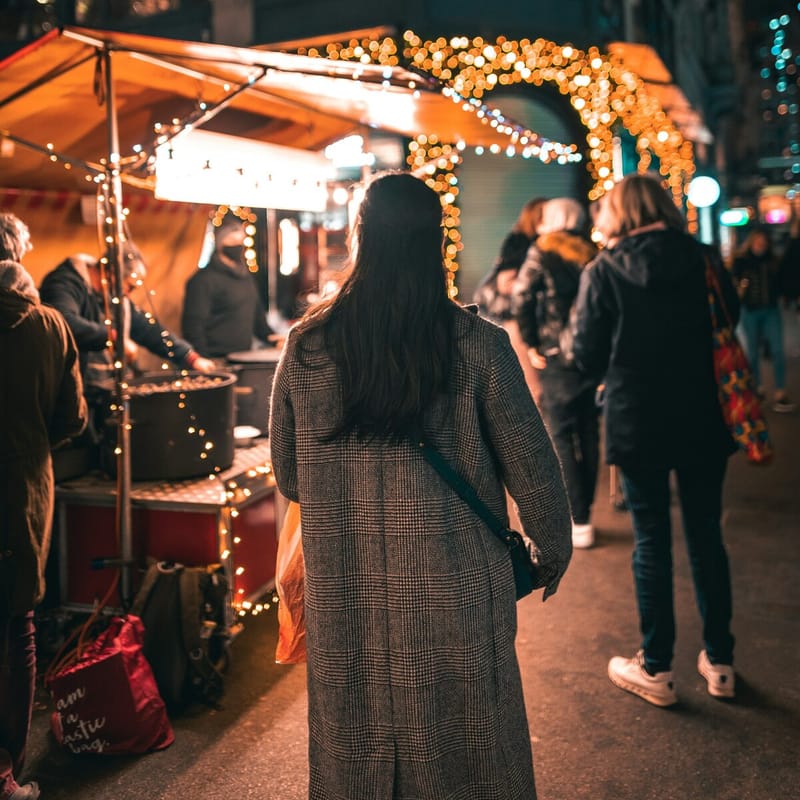A woman in a long tweed coat walks between lit market stalls at night, following friends under an arch of string lights.