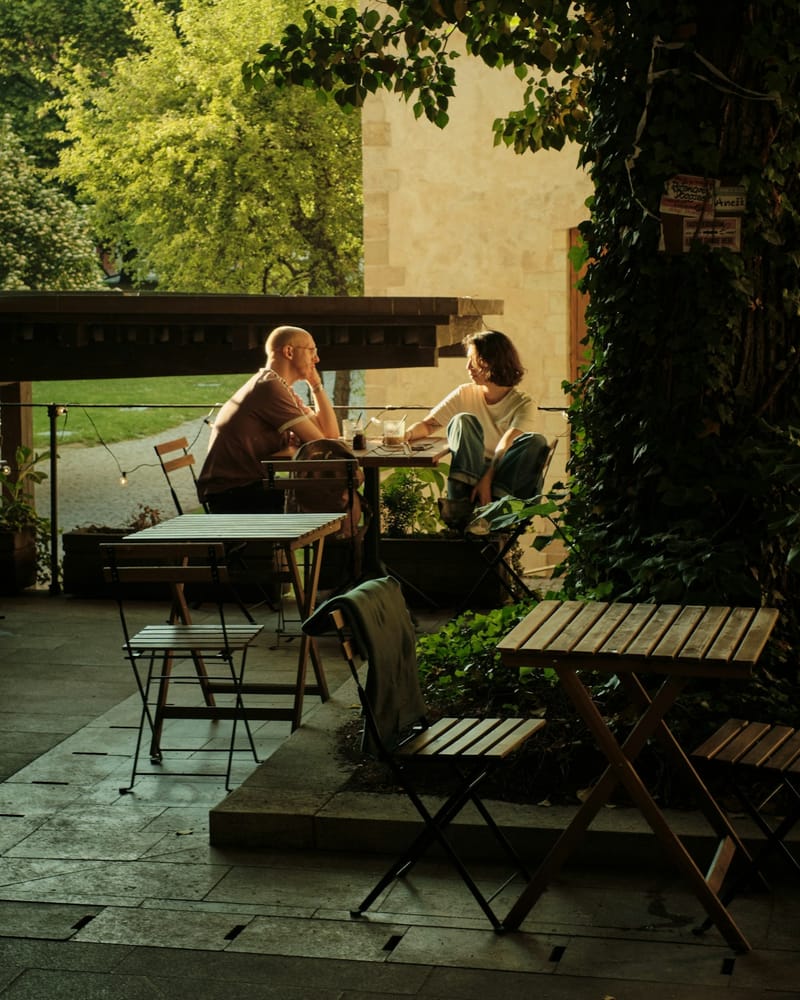 Two people talking across a small table on a café patio, lit by low golden evening sun beside an ivy-covered tree.