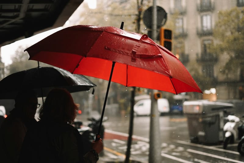 A red umbrella held over pedestrians on a rainy city street, raindrops beading on the canopy above a blurred road.