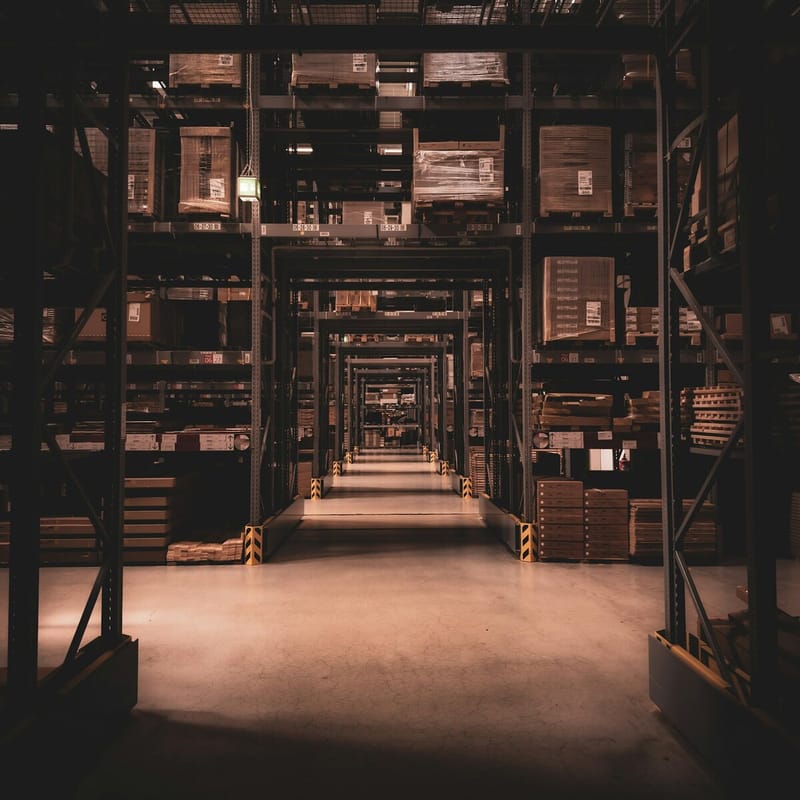 A long warehouse aisle between tall steel racks stacked with boxes, its concrete floor lit by low warm light.