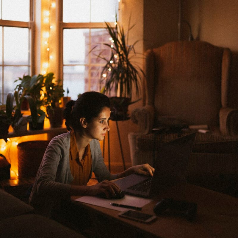 A young woman works on a laptop at a low table in a dim living room, lit by fairy lights strung along the window.