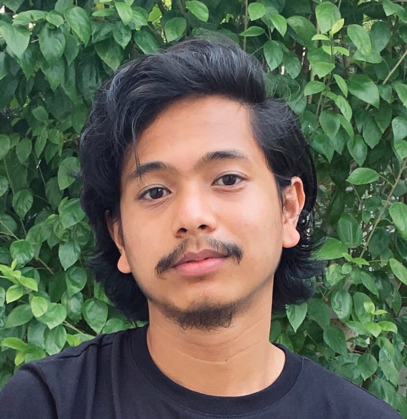 Redza Remee, writer at Carraway & Willow, with dark wavy hair and a goatee, in a black t-shirt before green leaves.